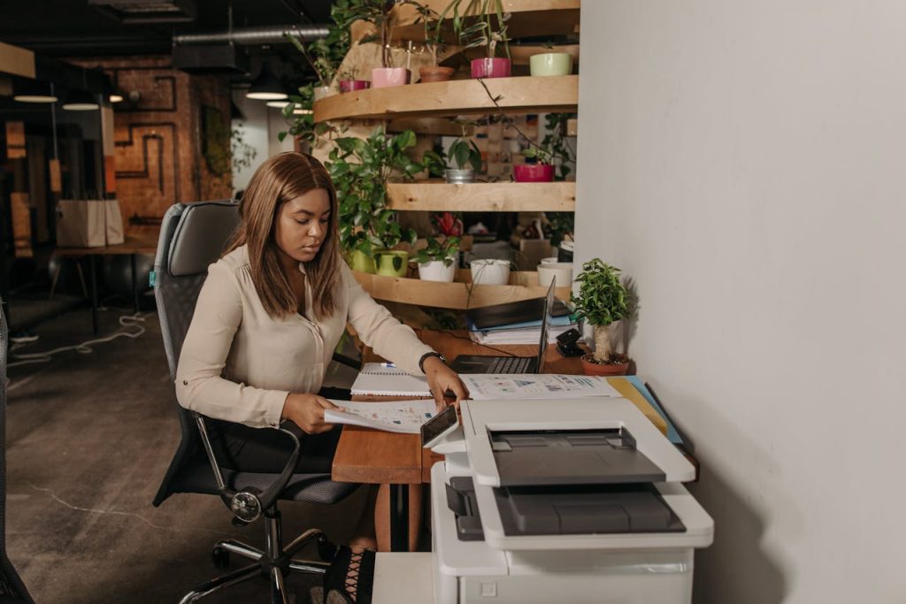 A focused woman works at a desk in a modern office environment, surrounded by plants.